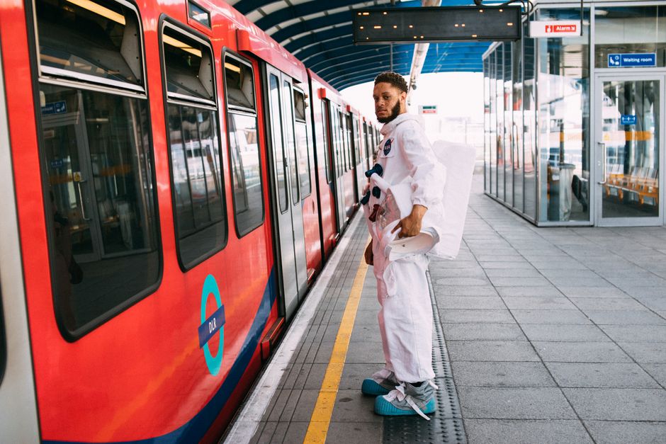 A man in a spacesuit stands by a train at a London station, merging sci-fi with urban comm