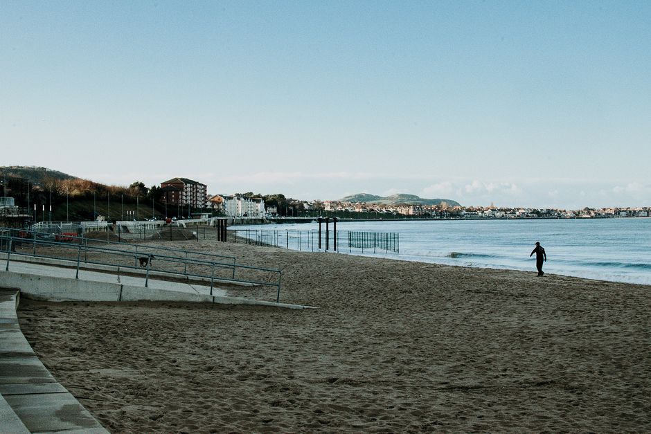 A tranquil beach view with a lone person walking on the sandy shore near buildings.