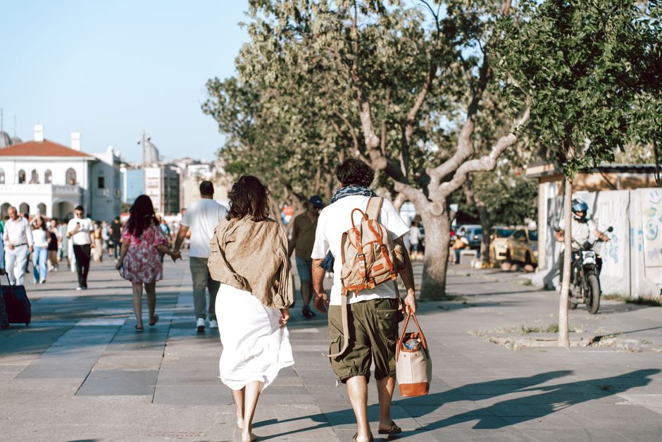 A couple strolls down a bustling city street surrounded by lush trees and vibrant urban life.