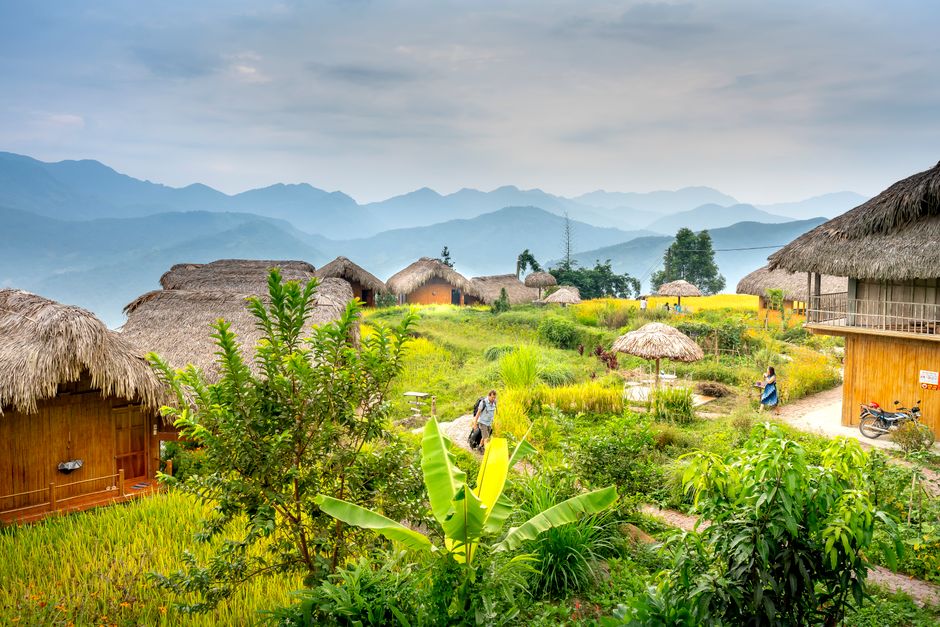 Anonymous tourists strolling in remote exotic hotel with bungalows in green grassy hills under bright sky in daytime