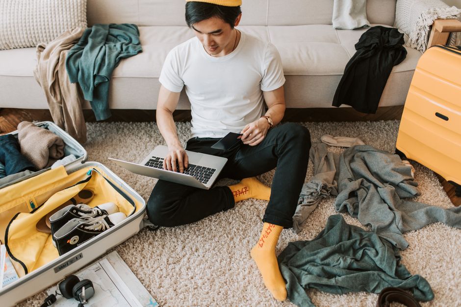 Man multitasking by packing luggage while working on a laptop in living room.