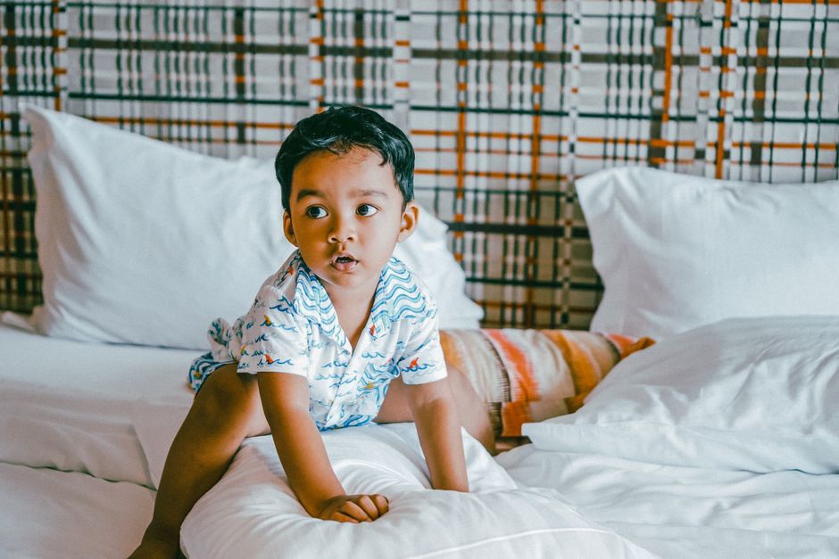 Adorable child sitting on a bed surrounded by pillows in a cozy indoor setting.