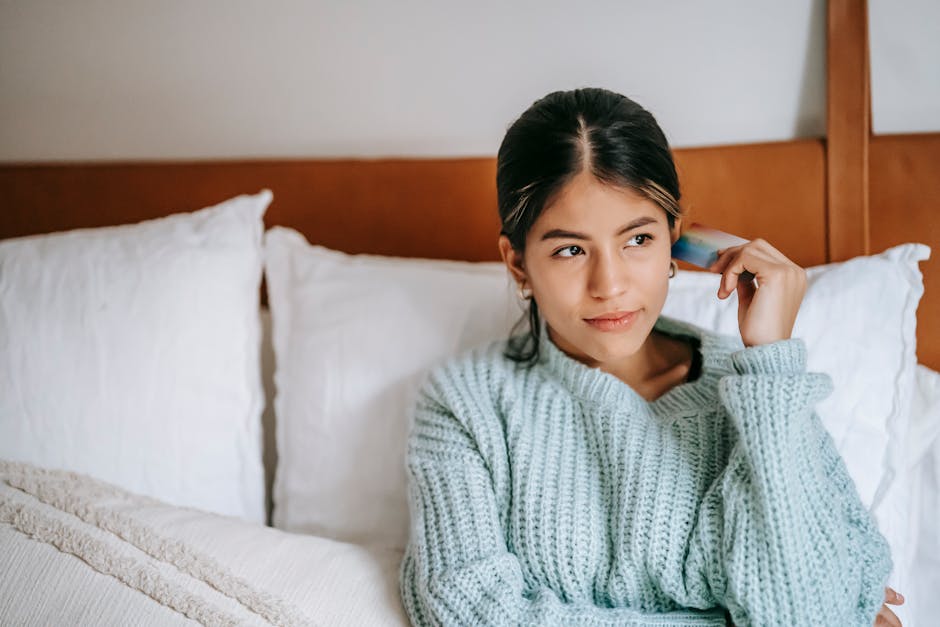 Young contemplative ethnic female with plastic card sitting on soft bed while looking away in house