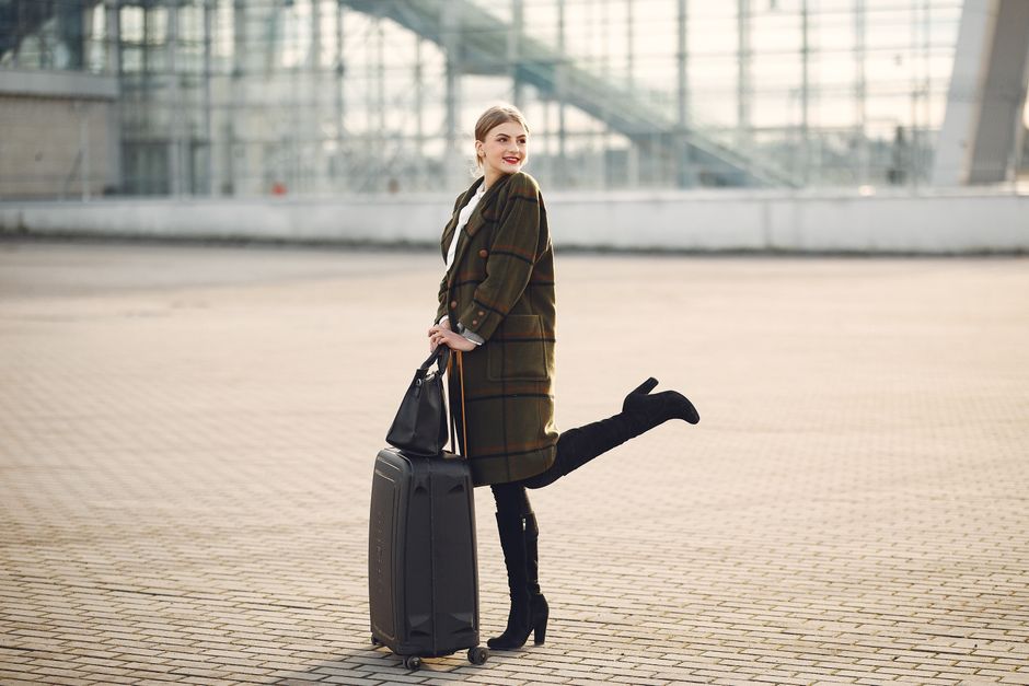 Full body of stylish female tourist in warm clothes and high heeled boots standing on background of blurred airport terminal with luggage and ladies bag