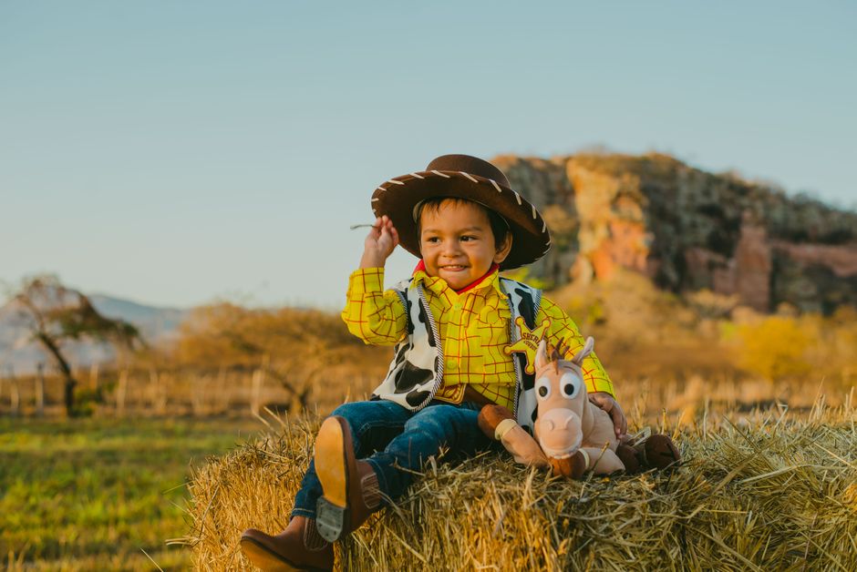 Cute child dressed as a cowboy sitting on hay with a stuffed toy, enjoying a sunny outdoor scene.