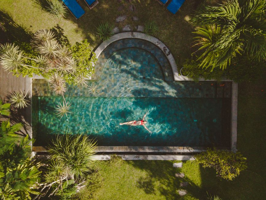 Aerial shot of a woman in a red swimsuit floating in a tropical outdoor swimming pool surrounded by lush greenery.