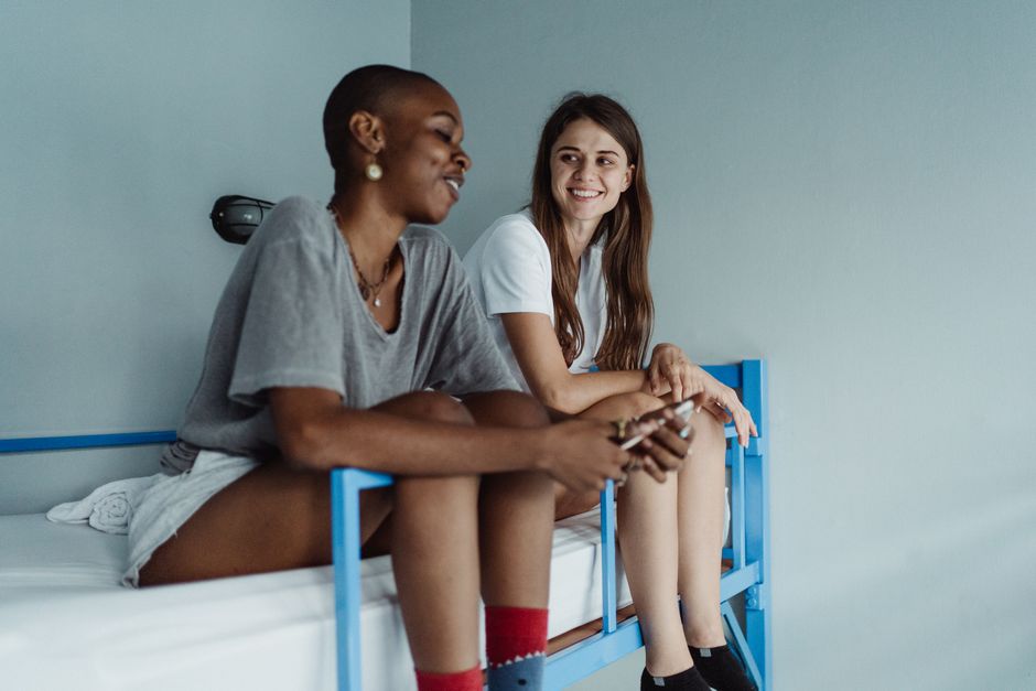 Two women smiling and conversing on a bunk bed indoors.