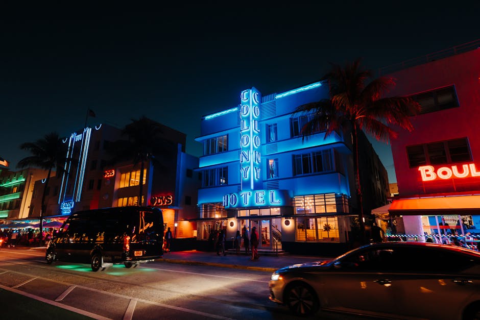 Glowing neon signs illuminate the Colony Hotel at night, showcasing vibrant nightlife in an Art Deco district.