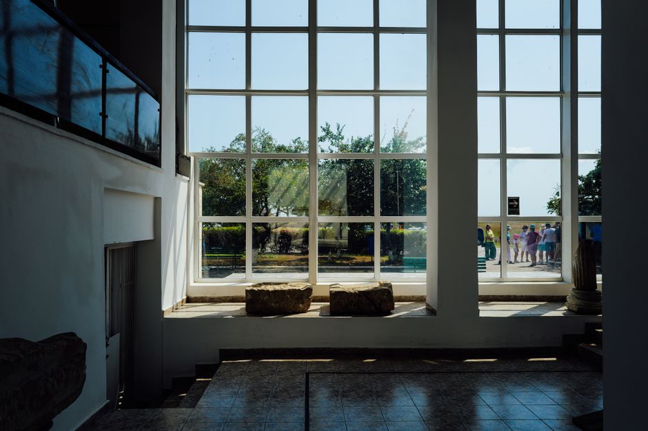 Interior view with large window overlooking people and greenery on a sunny day.