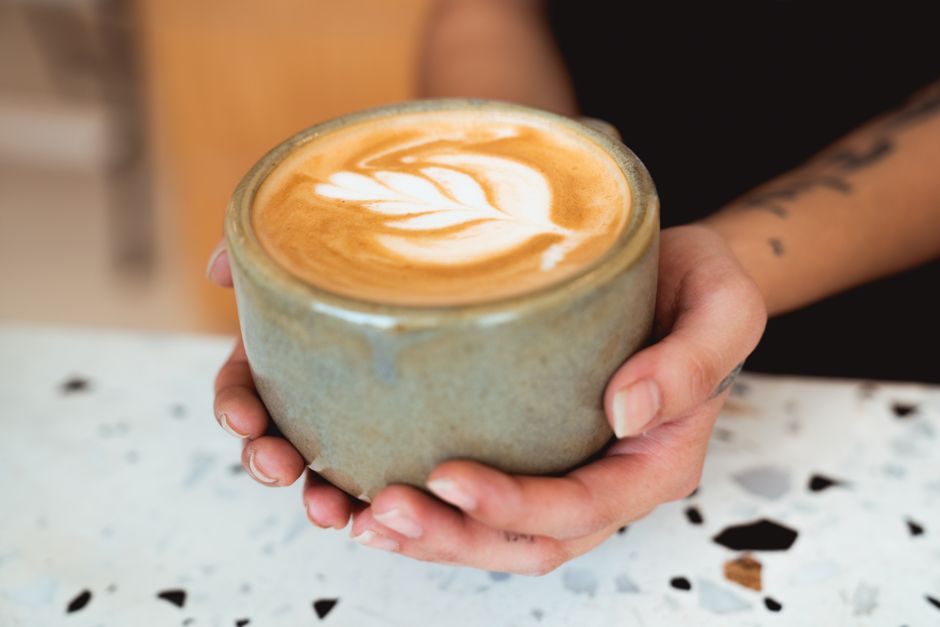 A close-up of a latte with leaf art in a ceramic mug, held by hands.