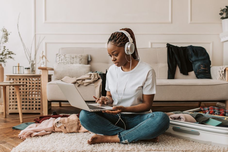 A young woman sits on the floor using a laptop and headphones, amidst clothes and a suitcase.