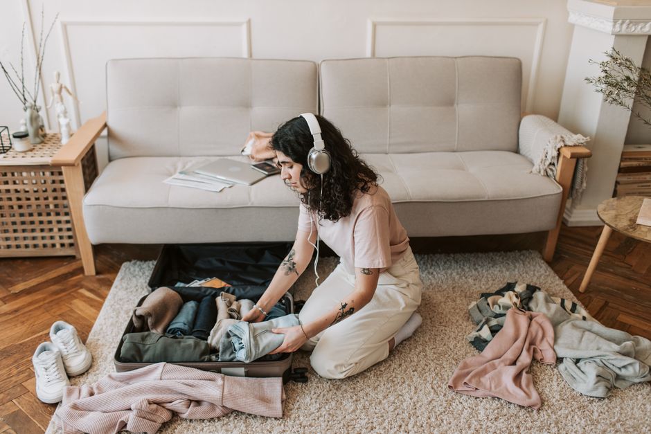 Woman packing suitcase on living room floor, wearing headphones, preparing for a trip.
