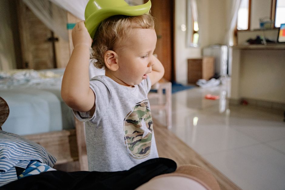 A young boy playing indoors, putting on a green plastic hat in a cozy bedroom setting.