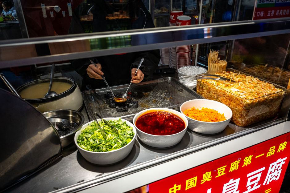 Street food vendor preparing a spicy tofu dish with various garnishes in an Asian market.