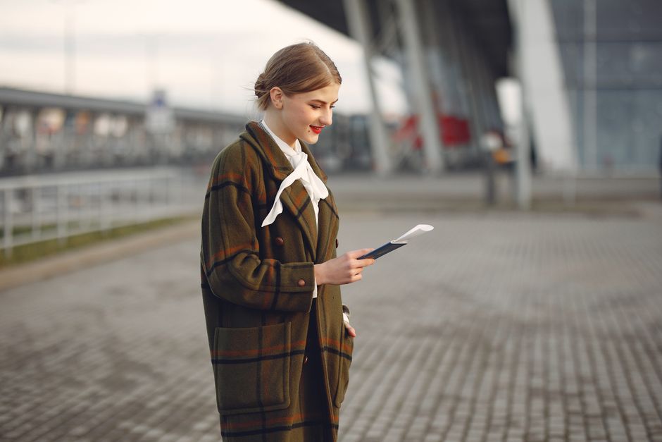 Smiling female passenger wearing trendy plaid coat and white blouse checking passport and ticket standing on pavement near contemporary building of airport