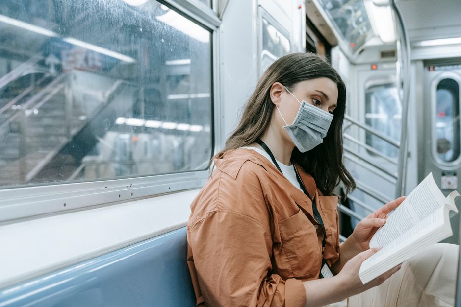 A woman in a face mask reads a book while commuting on a subway train.